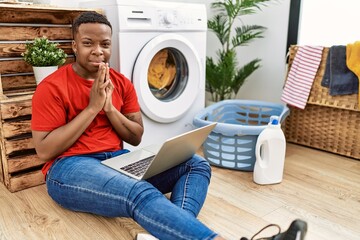 Poster - Young african man doing laundry and using computer praying with hands together asking for forgiveness smiling confident.