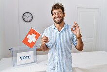 Young Handsome Man At Political Campaign Election Holding Switzerland Flag Doing Ok Sign With Fingers, Smiling Friendly Gesturing Excellent Symbol
