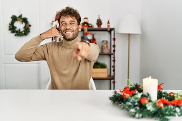 Wall Mural - Young handsome man with beard sitting on the table by christmas decoration smiling doing talking on the telephone gesture and pointing to you. call me.