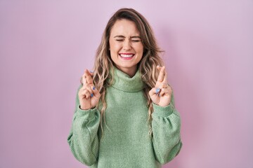 Sticker - Young caucasian woman standing over pink background gesturing finger crossed smiling with hope and eyes closed. luck and superstitious concept.