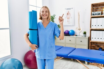 Poster - Young caucasian physiotherapist woman holding yoga mat smiling happy pointing with hand and finger to the side