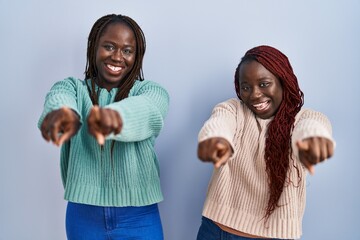 Sticker - Two african woman standing over blue background pointing to you and the camera with fingers, smiling positive and cheerful