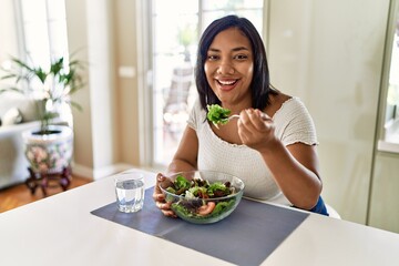 Hispanic brunette woman eating green salad at the kitchen