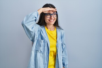 Canvas Print - Young hispanic woman standing over blue background very happy and smiling looking far away with hand over head. searching concept.