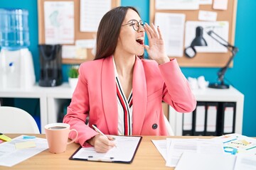 Canvas Print - Young hispanic woman working at the office wearing glasses shouting and screaming loud to side with hand on mouth. communication concept.