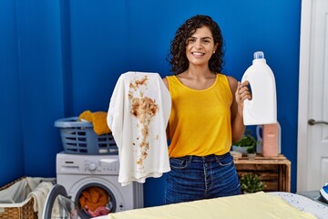 Poster - Young latin woman holding dirty t shirt and detergent bottle at laundry room