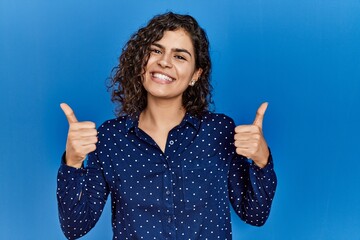 Poster - Young brunette woman with curly hair wearing casual clothes over blue background success sign doing positive gesture with hand, thumbs up smiling and happy. cheerful expression and winner gesture.