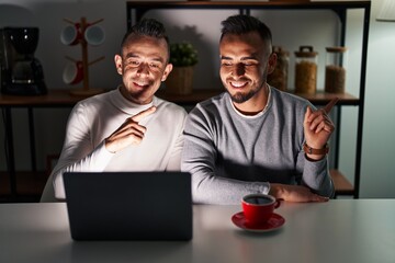 Poster - Homosexual couple using computer laptop with a big smile on face, pointing with hand finger to the side looking at the camera.