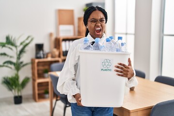 Poster - African american woman working at the office holding plastic bottle for recycling angry and mad screaming frustrated and furious, shouting with anger. rage and aggressive concept.