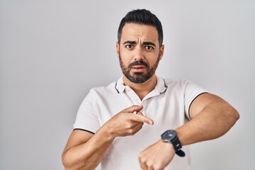 Poster - Young hispanic man with beard wearing casual clothes over white background in hurry pointing to watch time, impatience, upset and angry for deadline delay