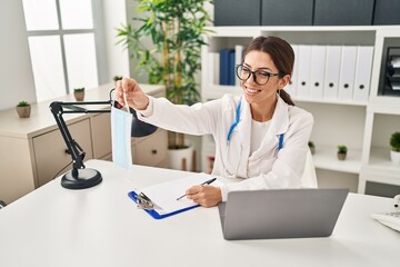 Canvas Print - Young hispanic woman wearing doctor uniform holding medical mask at clinic