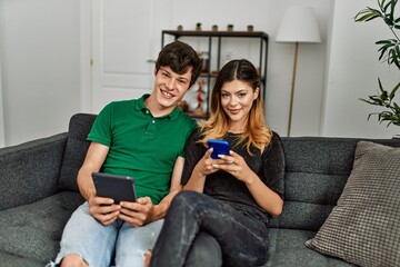 Poster - Young caucasian couple smiling happy using touchpad and smartphone at home.