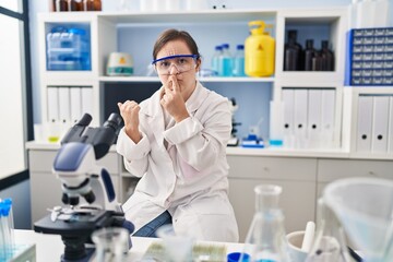 Poster - Hispanic girl with down syndrome working at scientist laboratory asking to be quiet with finger on lips pointing with hand to the side. silence and secret concept.