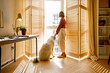 © rh2010 - Young woman stands with her dog by the window blinds and looks away in cozy and sunny living room of modern apartment in beige tones. Quarantine, loneliness and life at home concept