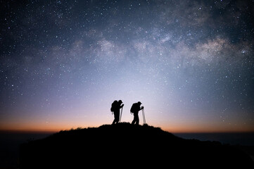 Wall Mural - Silhouette of young couple hiker were standing at the top of the mountain looking at the stars and Milky Way over the twilight sky. Both of them were happy and free to travel.