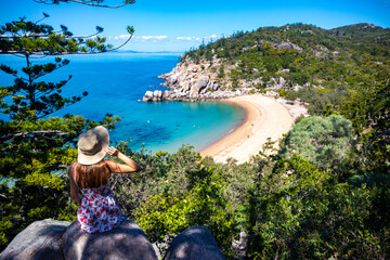 Wall Mural - beautiful long-haired girl in dress and hat sits on top of mountain overlooking paradise beach on magnetic island; beach with massive rocks and turquoise water; vacationing on magnetic island, queensl