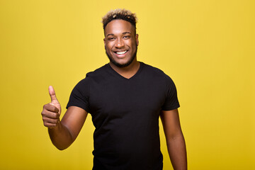 Wall Mural - Dark-skinned, cheerful young man points his thumbs up while standing on a yellow background. A satisfied young man shows a positive hand gesture, he is satisfied with everything. 