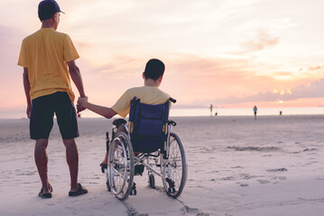 Behind of young man with disability holding hand father or volunteer or caregiver on the sea beach at sunset, Travel and vacation in summer, Positive photos give life power, mental health concept.