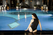 Woman in black swimsuit sitting relaxed by the swimming pool indoors, spending holidays at spa resort. View from the backside