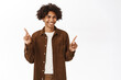 Portrait of smiling gen z guy with afro hair, pointing fingers sideways, showing left and right side, logo or product banners, give choice, standing over white background