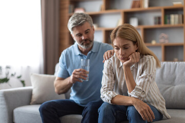 Wall Mural - Worrying middle aged european man gives glass of water and calms depressed woman on sofa in living room