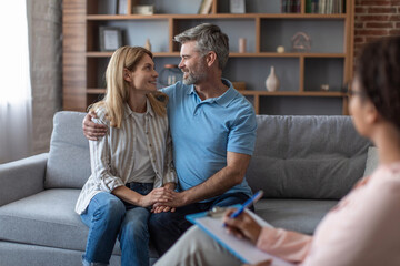 Wall Mural - Cheerful black young doctor in glasses looks at middle aged european couple hugging on sofa in clinic