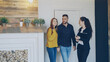 Cheerful female realtor is meeting beautiful young couple in new house opening door showing documents and talking to clients. Happy people and selling real estate concept.