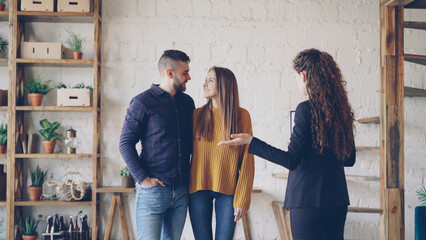 Wall Mural - Cheerful young people are talking with housing agent and hugging standing inside loft style house with beautiful interior. Purchasing property and accommodation concept.