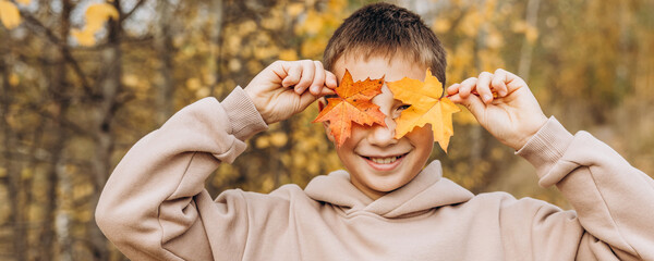 Teenager boy hiding his eyes behind maple leaves. Child holding yellow autumn leaves in his hands. Teen having fun on walking in autumn park. Selective focus