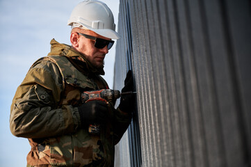 Canvas Print - Male builder installing black corrugated iron sheet used as facade of future cottage. Man worker building wooden frame house. Carpentry and construction concept.