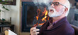 © Solid photos - Smiling senior man smoking pipe and reading book beside fireplace in winter