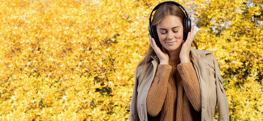 Happy woman standing on yellow autumn leaves background listening to music in headphones. Happy young girl listening to a podcast. Banner. Lifestyle, autumn fashion, beauty