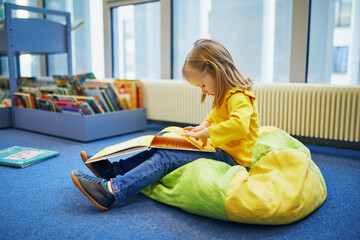 Wall Mural - 4 year old girl sitting on the floor in municipal library and reading a book