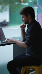 Young man working remotely at co working office by window overlooking city street sidewalk. Thoughtful person using laptop computer in Vertical Video