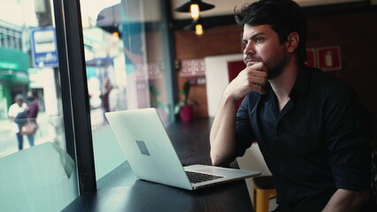 Concerned pensive young entrepreneur man in front of laptop computer seated by window at cafe co working remote workplace