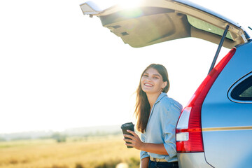 Wall Mural - happy young woman holding coffee cup and sitting on the open trunk of her car in a wheat field at sunset