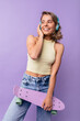 © Look! - Cute young caucasian girl listens to music through headphones looking away on purple background. Blonde smiles, holds skateboard, wears top and jeans. Concept of leisure, use