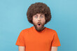 © khosrork - Portrait of amazed man with Afro hairstyle wearing orange T-shirt standing with mouth open in surprise, has shocked expression, hears unbelievable news. Indoor studio shot isolated on blue background.