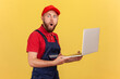 © khosrork - Side view of amazed surprised handyman standing laptop in hands and working online, looking at camera with big eyes, wearing uniform and red cap. Indoor studio shot isolated on yellow background.