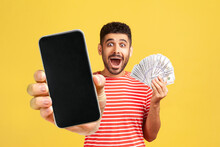 Surprised Happy Man With Beard In Red T-shirt, Looking With Big Amazed Eyes And Open Mouth, Holding Money And Showing Mobile Phone With Empty Screen. Indoor Studio Shot Isolated On Yellow Background.