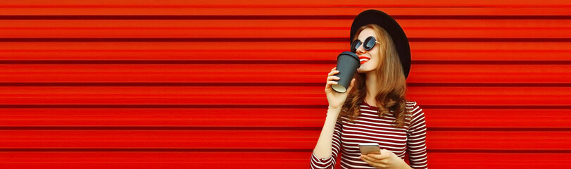 Portrait of happy smiling woman with smartphone drinking coffee looking away wearing black round hat on red background, blank copy space for advertising text