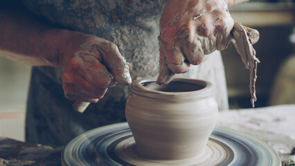 Elderly bearded craftsman is creating ceramic jar on potter's wheel using professional tools. Workplace with handmade pots, beautiful vases and figures in background.