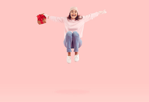 Happy cheerful excited smiling pretty woman in winter hat holding gift box tied with red bow, jumping high in mid air on pastel pink color background. Christmas presents and holiday sale concept