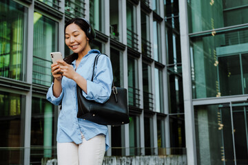 Asian brunette woman using cellphone while walking on city street