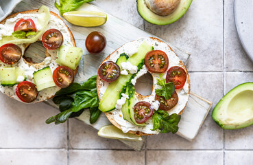 Wall Mural - Bagels sandwich topped with cream cheese, farm cottage cheese, avocado, tomatoes, cucumbers and salad, grey tilled background. Healthy breakfast food.