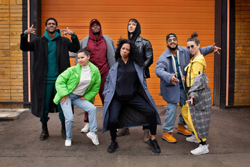 Multiracial group of young hipsters dance together on city street against orange colourful background. Diverse Stylish people outdoor, youth culture