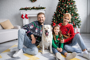 Wall Mural - Family in christmas sweaters and jeans looking at camera near labrador at home