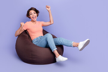 Full length photo of excited lucky lady wear striped t-shirt sitting bean bag rising fists empty space isolated violet color background