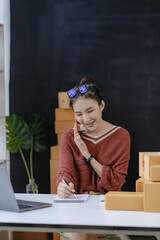 Wall Mural - An Asian businesswoman works online shopping at her store. A young female seller prepares a parcel box to send to a concept customer. online selling ecommerce vertical image