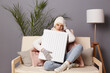 © sementsova321 - Full length portrait of woman wearing cap, gloves, scarf and coat sitting with crossed legs on sofa in home interior and holding heater for warming up.